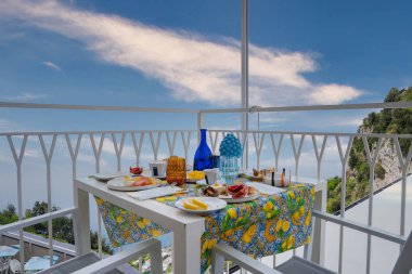 Limon desenli masa örtüsü, kahvaltı malzemeleri ve modern mobilyaların bulunduğu bir balkon. Yeşillik ve berrak gökyüzü ile Tyrhenian Denizi manzaralı..
