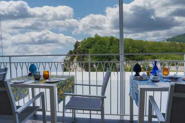 İtalya 'nın Amalfi kentindeki bulutlu bir gökyüzünün altında yeşil tepelere bakan korkuluk çerçeveli, renkli masa ayarlarıyla bir açık hava yemek alanı..