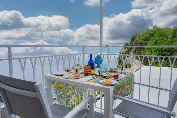 Limon desenli masa örtüsü, kahvaltı malzemeleri ve dekoratif cam kaplama, yeşil kayalıklar ve Amalfi 'de mavi gökyüzü olan bir balkon yemek takımı..