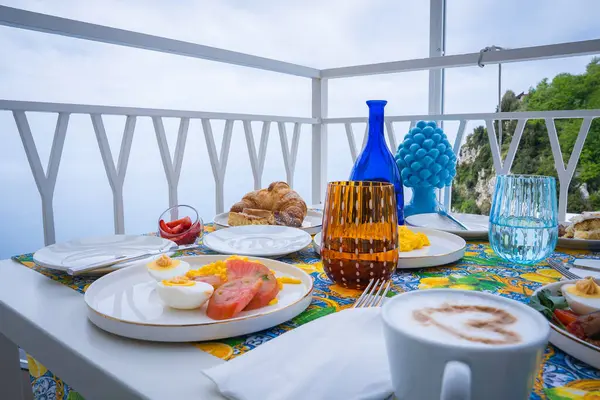 Yumurta, somon, kruvasan ve meyve içeren, Tyrhenian Denizi ve yeşilliğine bakan, mavi seramik dekorlu bir balkon kahvaltısı..
