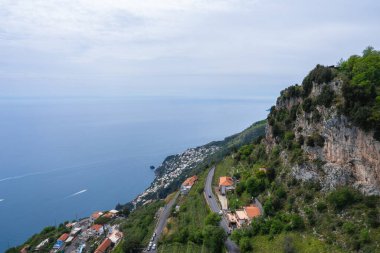 İtalya 'daki Amalfi Sahili' nde kayalıklar, turuncu çatılı teraslı binalar, dolambaçlı bir yol, yemyeşil bir yol ve Tyrhenian Denizi 'nde bir tekne yer alıyor..
