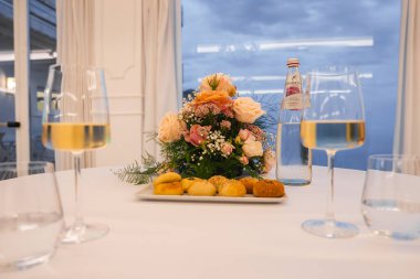 İçinde pembe ve şeftali gülleri, ekmek, beyaz şarap ve cam eşyalar olan bir masa seti. Büyük pencereler bulutlu bir gökyüzü ve İtalya 'nın Amalfi kentindeki Tyrhenian Denizi' ni gösteriyor.
