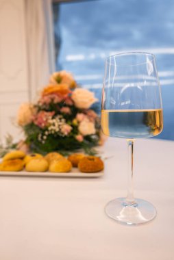 Ön planda pastalar ve pastel güller olan beyaz bir masa örtüsü üzerinde bir bardak beyaz şarap. Amalfi 'deki bir pencereden bulutlu bir gökyüzü görülebilir.