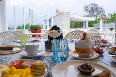 Örtülü kumaş üzerinde yumurta, hamur işi ve meyve olan bir kahvaltı masası. Terasta beyaz sandalyeler, cam korkuluklar, taş duvar ve yeşillik yer alıyor..