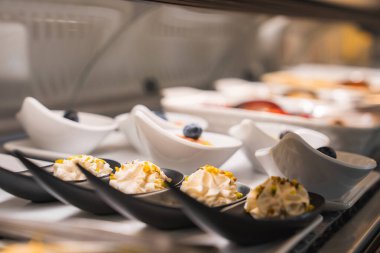 Ön planda krem şantili ve şam fıstıklı küçük siyah kaşıklar, rafine edilmiş Amalfi soslu beyaz kaseler ve yaban mersini..