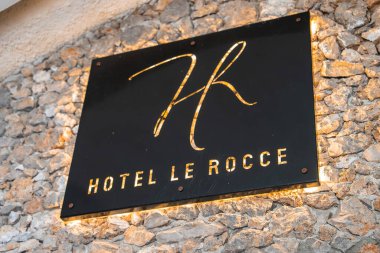 Altın yazılı siyah tabela ve Hotel Le Rocce için stilize logo, kalın ışıklandırma ile dokulu taş bir duvara monte.