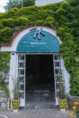 İtalya, Amalfi 'de bir otele giriş, sarmaşık kaplı taş bir duvar, deniz mavisi çokdilli hoş geldin işareti ve bitkilerle dekoratif seramik kaplar.