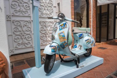 Capri temalı çıkartmalar ve kartpostallarla süslenmiş açık mavi bir scooter Capri, İtalya 'da beyaz işlenmiş demir bir kapı ve tuğla çerçeveli bir mağazanın önünde duruyor..