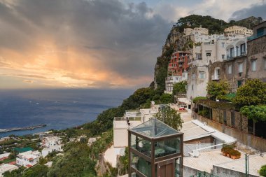 Günbatımında İtalya 'daki Capri Adası' nda renkli kayalık binalar, cam bir asansör, Akdeniz, botlar ve yemyeşil yeşillikler yer alıyor..