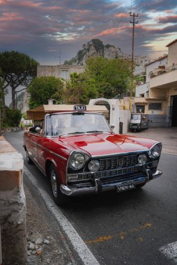 Krom aksanlı ve yuvarlak farlı üstü açık kırmızı bir taksi, İtalya 'nın Capri şehrinde dar bir sokağa park edilmiş, arka planda kayalıklar ve binalar var..