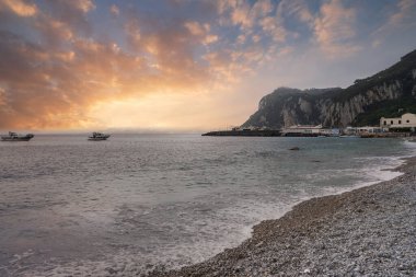 İtalya, Capri Adası 'ndaki sahil manzarası, çakıl taşı plajı, sakin suları, tekneleri, yeşillikli kayalık bir uçurumu ve turuncu gökyüzünün altında gün batımında bir liman..