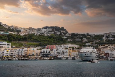 İtalya 'nın Capri kentindeki Marina Grande' de, sıcak bir günbatımı gökyüzü altında renkli deniz manzaralı binalar, büyük bir yat, küçük tekneler ve yemyeşil tepeler yer alıyor..