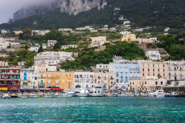 Capri, İtalya 'daki Marina Grande' de canlı binalar, kenetlenmiş tekneler, turkuaz su, yemyeşil tepeler, beyaz villalar ve sisli kayalıklar yer alıyor..