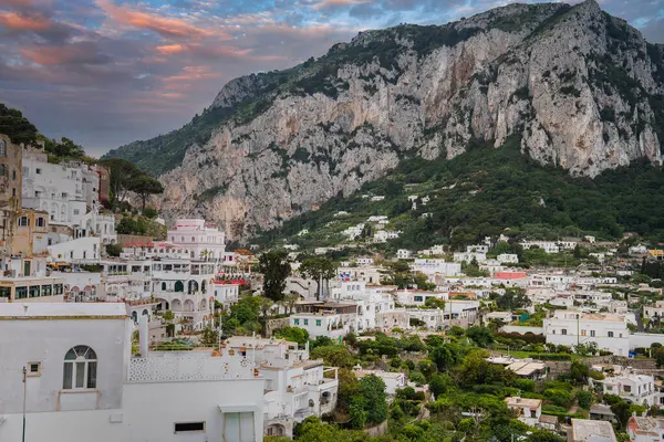 Capri, İtalya 'da bir yamaçta beyazlatılmış binalar, engebeli bir kireçtaşı dağı ve gün batımında pembe ve turuncu gökyüzü arka planda..