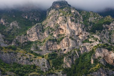 Yeşil benekli sarp kayalıklar, kaya yüzeyine oyulmuş kemerli tüneller ve Amalfi kıyısında Positano yakınlarında alçak asılı bulutlar..