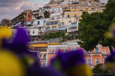Positano, İtalya manzarası, sarp arazi üzerindeki pastel renkli binalar, bir ağaç tarafından çerçevelenmiş ve bulanık çiçekler, pembe ve gri bir gökyüzü altında.