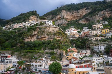 Positano, İtalya manzarası, sarp bir yamaçta canlı binalar, bitki örtüsü ile kayalık bir çıkıntı ve dramatik bir manzara yaratan bulutlu bir gökyüzü..