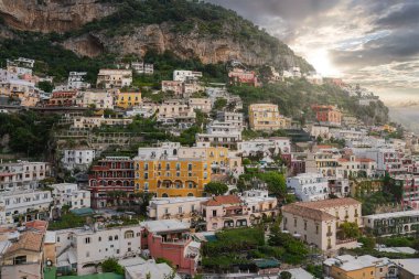 Positano, İtalya, sarp bir yamaçtan aşağı süzülen canlı binalar, yemyeşil, teraslı bahçeler, ve ılık bir günbatımı ışının altında kayalık bir uçurum..