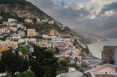 Amalfi kıyısındaki pozitano 'da istiflenmiş binalar, bir kilise kubbesi, yemyeşil alanlar, uçurumlar ve bulutlu bir gökyüzünün altındaki Akdeniz yer almaktadır..
