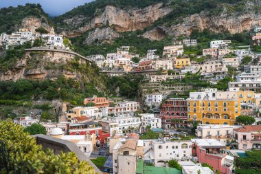 Yeşillik ve kayalık kayalıklarla çevrili sarı, kırmızı ve beyaz teraslı binaların bulunduğu Positano, İtalya manzarası.