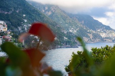 İtalya 'nın Positano şehrinin manzarası, gür kayalıklardaki renkli binalar, ön planda bulanık yeşillikler ve aşağıdaki Amalfi sahilinin mavi suları..