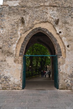 Yeşil metal kapılı Gotik tarzı taş kemer yolu ağaçlı ve patikalı bir bahçeye çıkıyor. İnsanlar yürür, ölçek ve aktivite ekler.