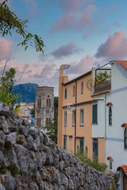 Ravello, İtalya 'da, etrafı pastel binalar, terracotta çatıları, yeşillik ve pembe mavi gökyüzü ile çevrili, kemerli pencereli taş bir çan kulesi bulunuyor..