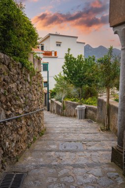 İtalya 'nın Ravello şehrinde, duvarlar ve yeşilliklerle çevrili, beyaz bir bina, ağaçlar, dağlar ve renkli bir günbatımı gökyüzü ile döşenmiş taşlı bir patika..