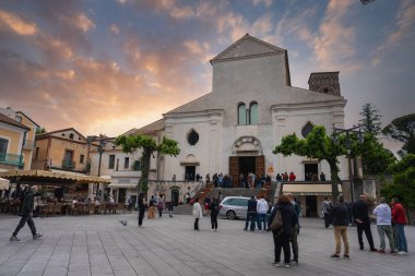 Ravello Katedrali 'nin beyaz cephesi ve çan kulesi Piazza Duomo' ya hükmediyor. İnsanlar toplanıyor ve kafeler sıcak bir günbatımı gökyüzünün altında..