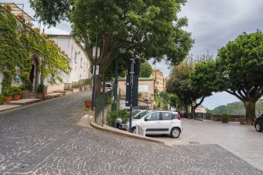 İtalya 'nın Ravello şehrinde kaldırımları, sarmaşık kaplı binaları, park edilmiş arabaları, yapraklı ağaçları ve bulutlu bir gökyüzünün altındaki tepe manzarası olan büyüleyici bir cadde..
