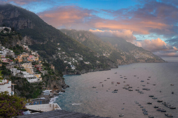 Живые здания Позитано каскадом спускаются по зеленым скалам к морю, с лодками на спокойных водах и закатом неба в розовых и оранжевых тонах.