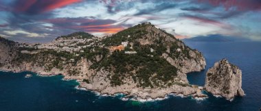 Capri 'nin engebeli kıyı şeridi kayalıklara, yemyeşil yeşilliklere, beyazlatılmış binalara ve Faraglioni kayalıklarına sahiptir. Pembe, mor ve mavi günbatımı gökyüzünün altında..