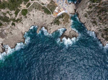 Capri Adası 'nın kayalık uçurumlarının derin mavi sularla buluştuğu hava manzarası. Dalgalar kıyı boyunca çarpar patikalar, yapılar ve dağınık şemsiyeler yakınında..