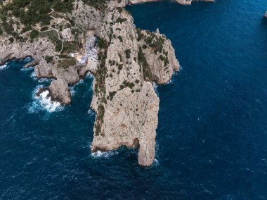 Capri Adası 'nın kireçtaşı uçurumları, derin mavi suları, küçük bir yapısı ve kenarına yakın renkli şemsiyeleri olan kayalık kıyı şeridinin dramatik hava manzarası..