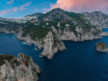 Capri Adası 'nın kıyı şeridinde kayalık kayalıklar, pürüzlü deniz yığınları, yemyeşil yemyeşil ve pembe ve mavi gökyüzünün altında beyaz yamaç binaları bulunur..