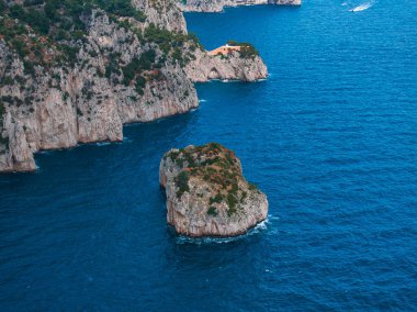 Yeşil renkli engebeli uçurumlar, Capri Adası 'nda mavi Akdeniz sularıyla buluşuyor. Bir kaya oluşumu ve bir uçurumun üzerindeki turuncu bina görsel ilgi ekliyor..