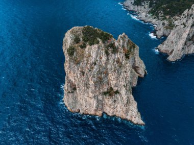 Amalfi Sahili yakınlarındaki mavi Akdeniz sularıyla çevrili engebeli kayalık, arka planda kayalıklar ve seyrek bitki örtüsü var..