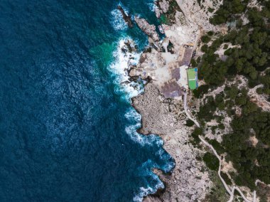 İtalya 'nın Capri Adası' nın hava manzarası kayalık uçurumları, derin mavi Akdeniz sularını, yeşil bir çatı binasını, patikaları ve yoğun yeşillikleri gösteriyor..