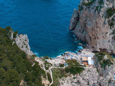 İtalya, Capri Adası 'ndaki sahil manzarası, sarp kayalıklar, canlı mavi sular, şemsiyeli küçük bir sahil ve dolambaçlı yollar boyunca yemyeşil..