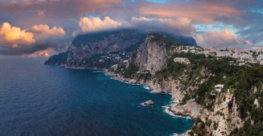 Kireçtaşı uçurumları, yemyeşil alanlar ve Capri Adası sahil şeridindeki beyaz binalar. Tyrhenian Denizi, turuncu ve pembe renkleriyle gün batımını yansıtır..