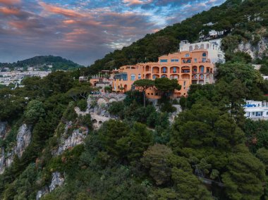 Kemerli pencere ve terasları olan turuncu bir villa Capri, İtalya 'da yeşil bir yamaçta, taş bir köprünün yanında ve pembe mavi gökyüzünün altındaki engebeli kayalıklarda bulunur..