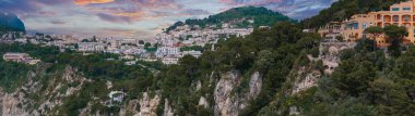 İtalya Capri Adası 'ndaki Capri kasabası, beyazlatılmış binalar, yemyeşil alanlar, kayalık uçurumlar ve gün batımında öne çıkan turuncu bir bina..