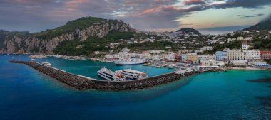 Capri Adası 'ndaki Marina Grande' de turkuaz sular, feribotlar, renkli binalar, yeşil tepeler, kayalık uçurumlar ve parçalı bulutlu bir akşam gökyüzü yer alıyor..