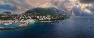 İtalya 'nın Capri Adası limanında yatlar, taştan bir iskele, beyazlatılmış tepe binaları, engebeli uçurumlar ve turkuaz suların üzerinde gün batımlı bir gökyüzü bulunur..