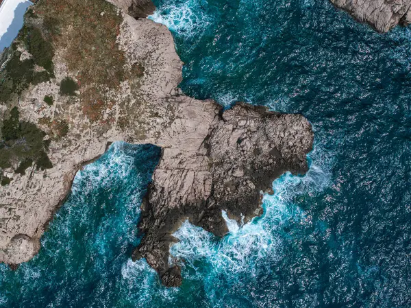 Canlı turkuaz ve derin mavi sularla çevrili engebeli kayalık kıyı şeridi. Dalgalar, hava görünümünde sivri kenarlara çarparlar.