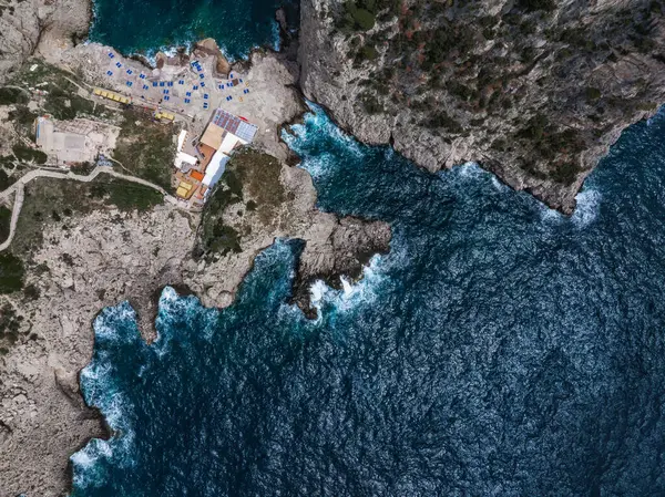İtalya, Capri Adası 'nın havadan görünüşü, kayalıkları, derin mavi denizi, çarpan dalgaları, binaların ve renkli plaj şemsiyelerinin olduğu gelişmiş bir alanı gösteriyor..