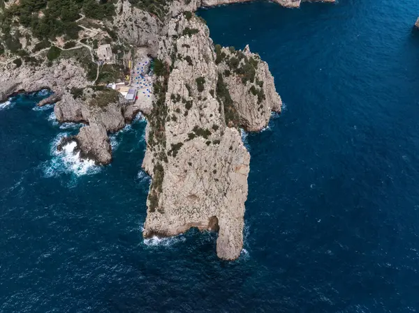 Capri Adası 'nın kireçtaşı uçurumları, derin mavi suları, küçük bir yapısı ve kenarına yakın renkli şemsiyeleri olan kayalık kıyı şeridinin dramatik hava manzarası..