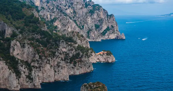 İtalya 'nın başkenti Capri Adası' nın engebeli kayalıkları, yeşil bir bina, turuncu bir çatı, derin mavi Akdeniz suları, uzak kayıklar ve parçalı bulutlu bir gökyüzü..