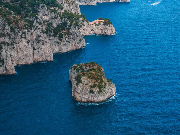 Yeşil renkli engebeli uçurumlar, Capri Adası 'nda mavi Akdeniz sularıyla buluşuyor. Bir kaya oluşumu ve bir uçurumun üzerindeki turuncu bina görsel ilgi ekliyor..