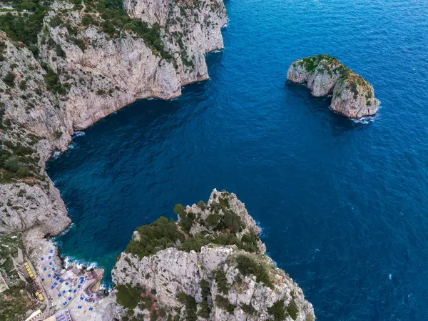 İtalya 'nın Capri Adası' nın havadan görünüşü, kireçtaşı kayalıkları, derin mavi bir koy, kaya oluşumları ve sol alt köşesinde şemsiyeler olan küçük bir plaj gösteriyor..